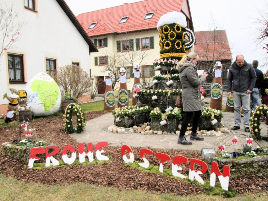 Besucher bewundern den Osterbrunnen. Im Vordergrund steckt im Blumenbeet der rot-weiße Schriftzug "Frohe Ostern".