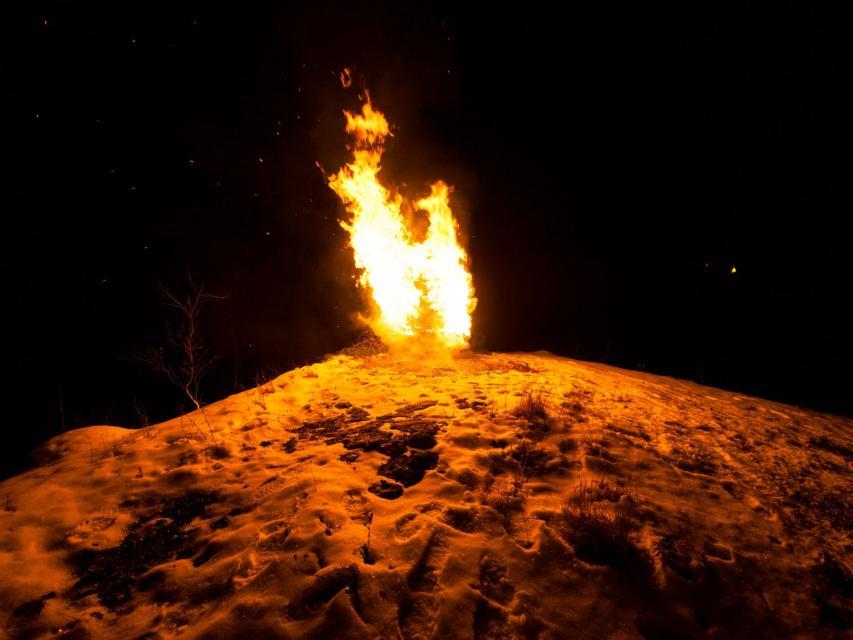 Auf einem schneebedeckten Hügel brennt ein Holzstapel. Die Flamme schlägt hoch in die Luft.
                 title=