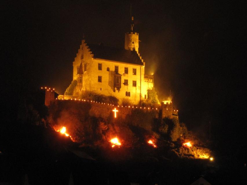 Nachtaufnahme der beleuchteten Burg. Unterhalb der Burgmauer brennen vier Holzfeuer. An der Burgmauer hängt ein beleuchtetes Kreuz.
                 title=