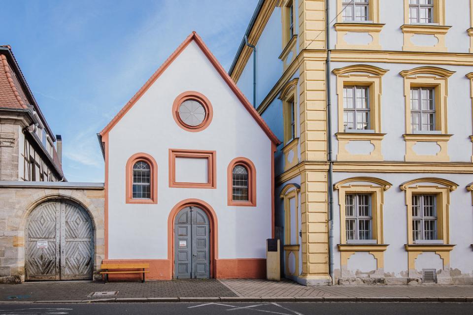 Weiß, mit terrakottafarbenen Umrahmungen der Fenster und der grauen Holztüre, steht die kleine Kapelle mitten in Forchheim.
                 title=