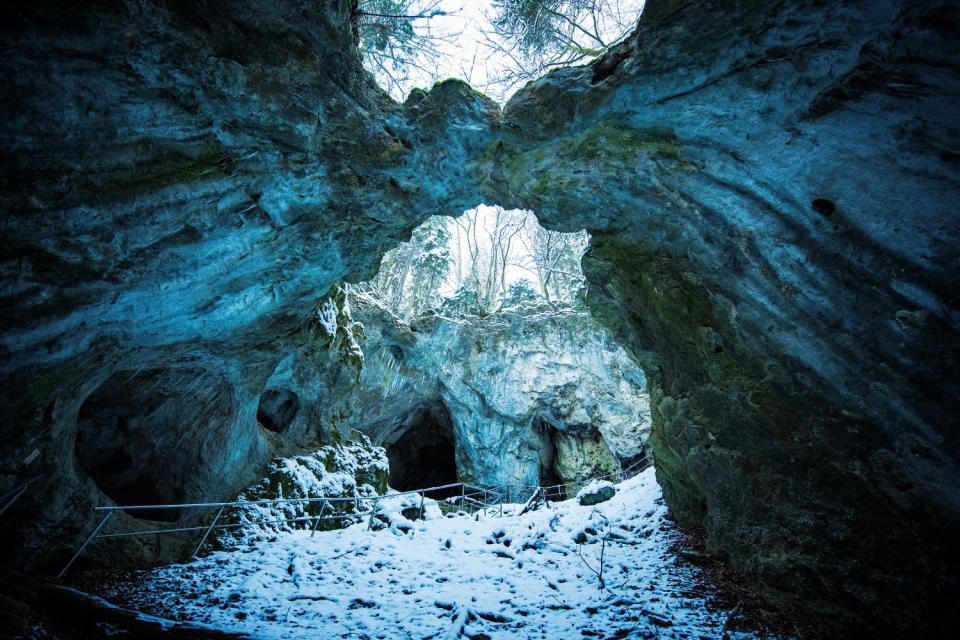 Verschneite Ansicht der Versturzhöhle. In der felsigen Decke sind zwei große Löcher, durch die man den Wald und Himmel sehen kann. Ein Weg mit Geländer führt bergauf durch die Höhle.