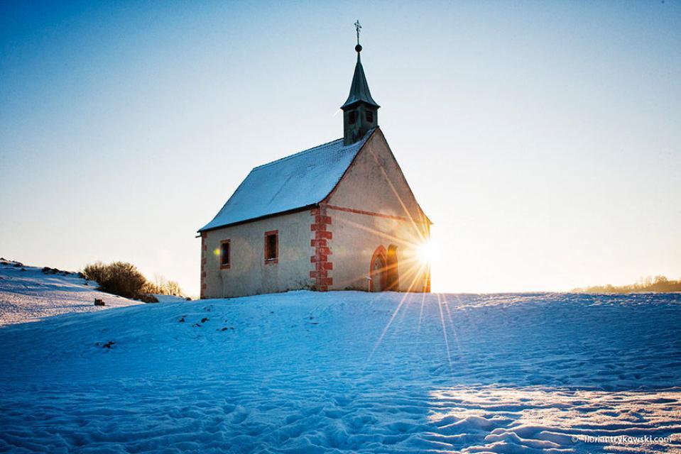 Auf einer schneebedeckten Wiese erhebt sich eine kleine Kapelle mit Glockenturm. Am Rand der Kapelle strahlt die Sonne im Sonnenuntergang.
