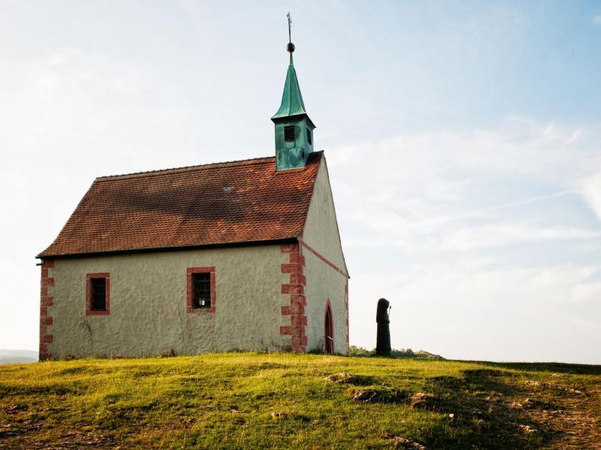 Auf einer grünen Wiese im Abendlicht erhebt sich die kleine Kapelle mit zwei Fenstern und einem Glockenturm. Davor steht eine Statue, von der nur der Umriss erkennbar ist.