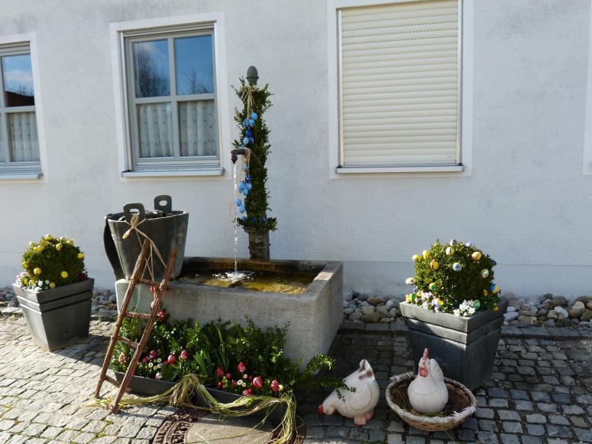 Vor einem Haus steht ein Betonbecken mit Wasserauslass. Grüne Girlanden, Butten, eine kleine Leiter und zwei Hühner zieren den Brunnen.