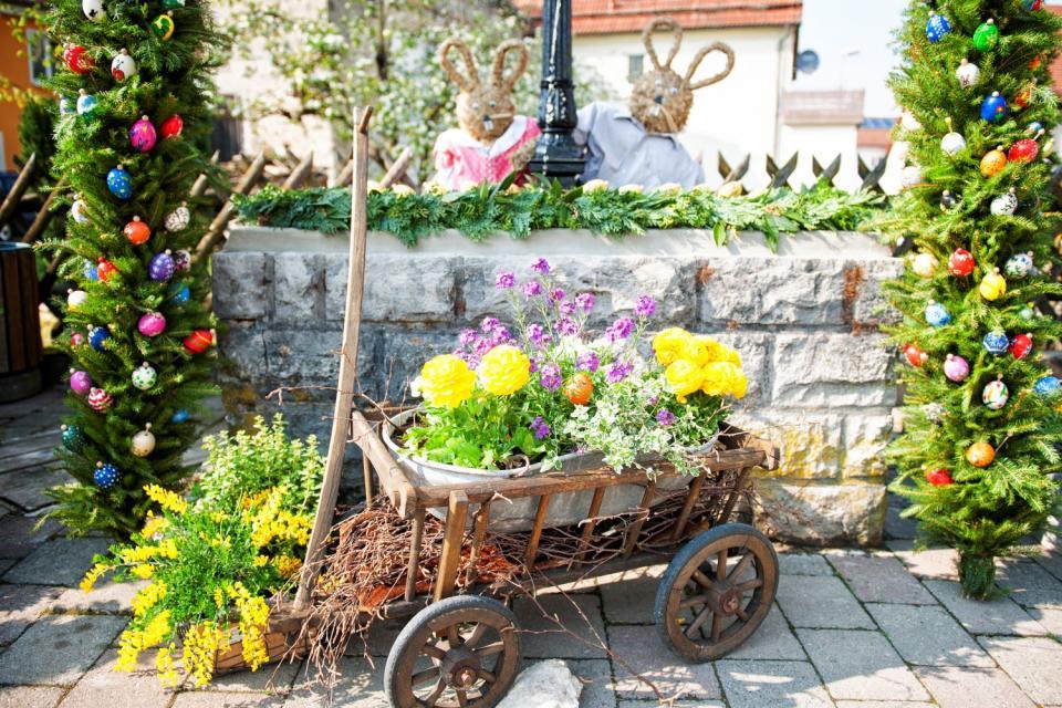 Vor dem steinernen Brunnen, der von grünen Girlanden mit Ostereiern umrahmt wird, steht ein kleiner Leiterwagen, in dem ein Blumentopf mit gelben und lila Blumen steht.