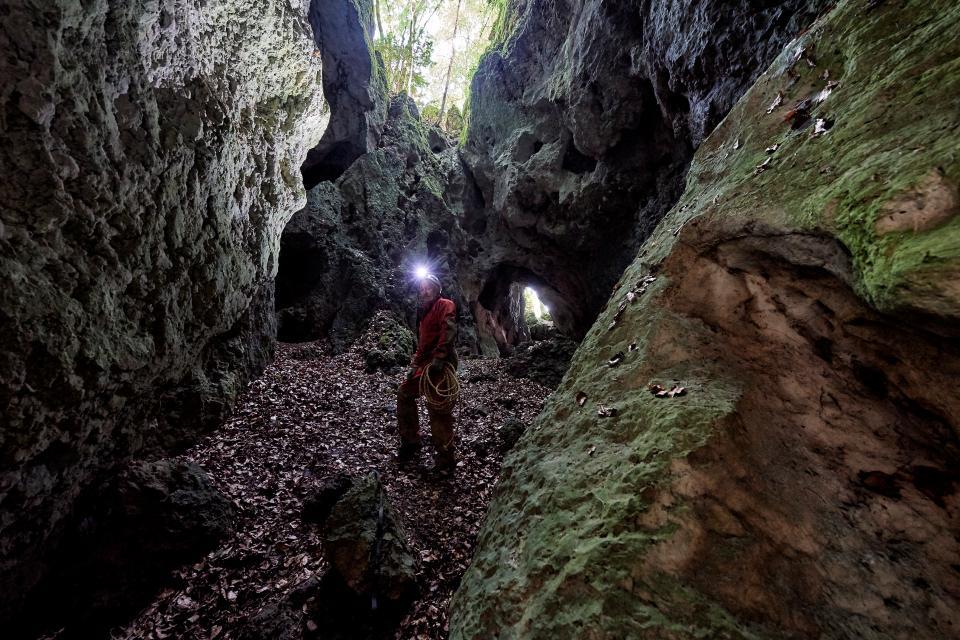 Zwischen aufragenden Felsbrocken steht auf belaubtem Waldboden ein Mann mit einer leuchtenden Stirnlampe und roten Overall.