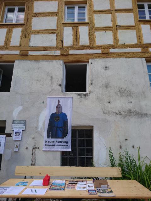 An der Fassade eines alten Fachwerkhauses hängt ein Banner mit einem Ritter. Vor dem Haus steht ein Biertisch, auf dem Prospekte ausliegen.