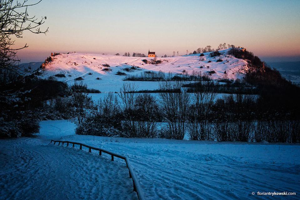 Blick über eine leicht schneebedeckte Landschaft. Im Hintergrund ehrebt sich ein Tafelberg in der Abendsonne. Darauf ist eine kleine Kapelle zu erkennen.
                 title=