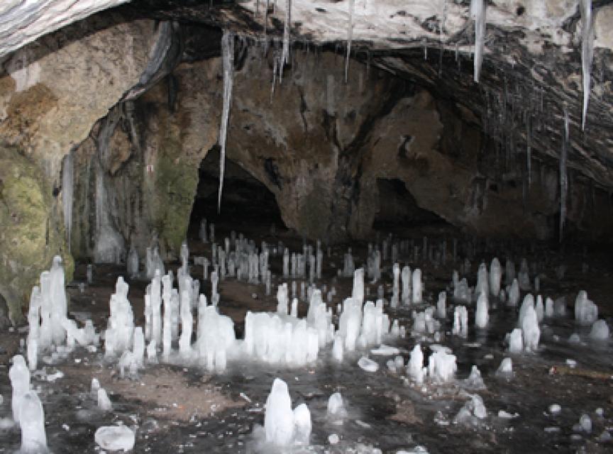 Im felsigen Höhleneingang ragen vom Boden Eiszapfen in die Höhe. Einige wenige Eiszapfen hängen von der Felsdecke.