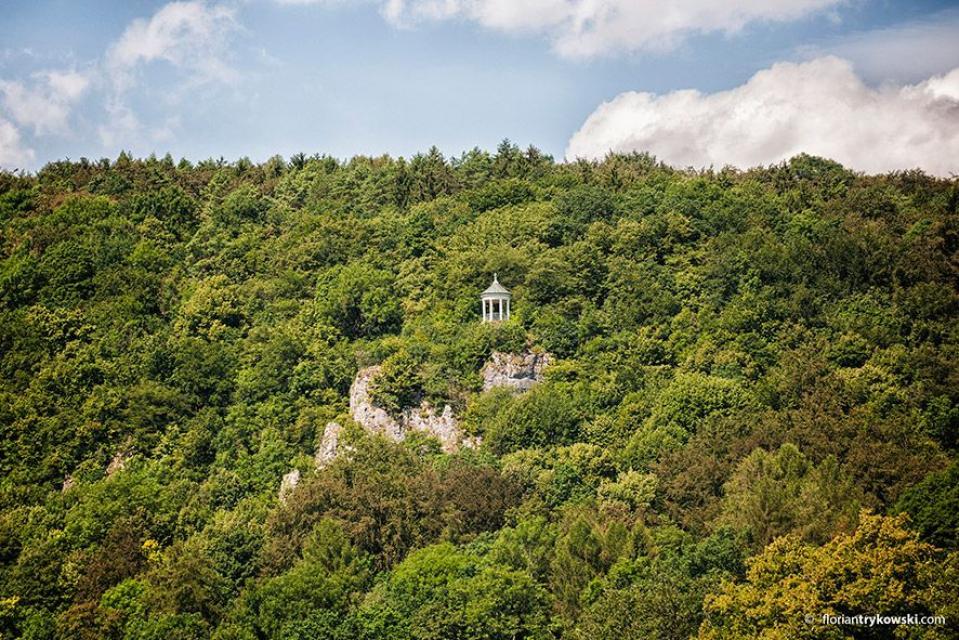Blick auf den abfallenden, bewaldeten Berghang. Auf halber Höhe ragen Felsen heraus, auf denen der Pavillon steht.