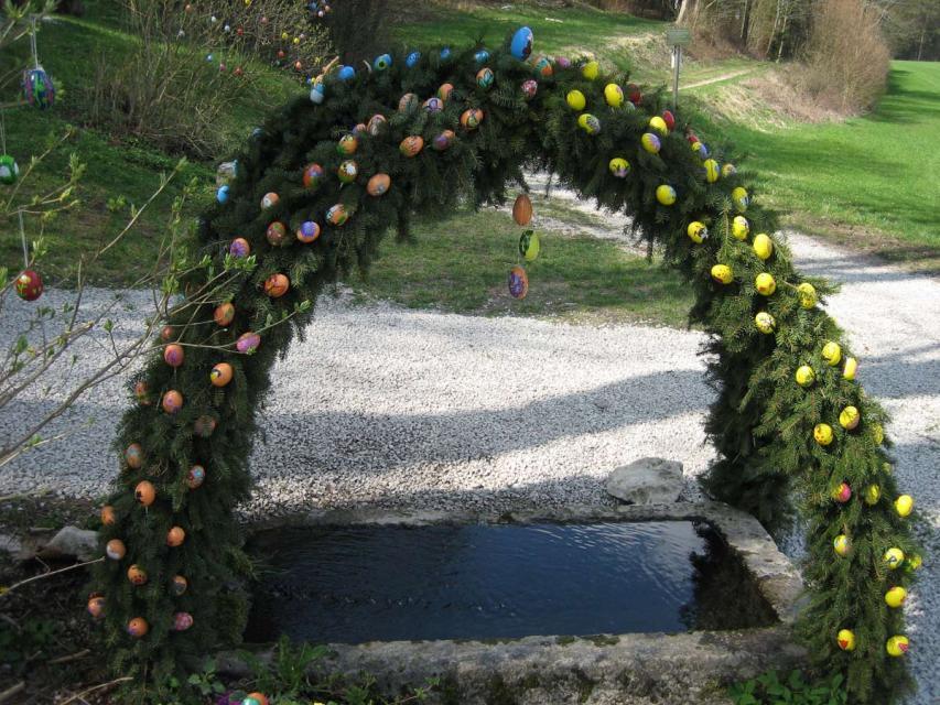 Über einem eckigen Brunnenbecken erhebt sich eine schlichte Krone aus grünen Girlanden mit bunten Eiern.