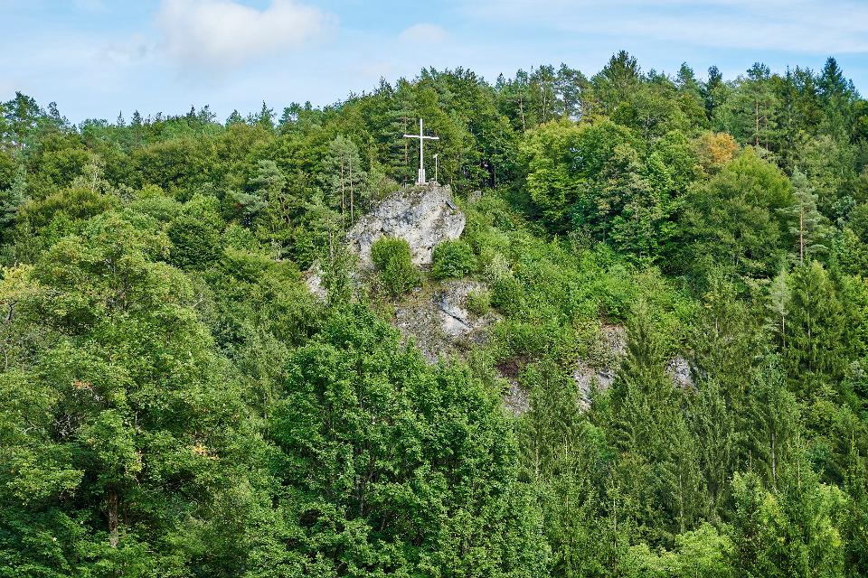 Mittig in einem steilen Waldhang, erhebt sich eine Felsen, auf dem eine Kreuz zu sehen ist.