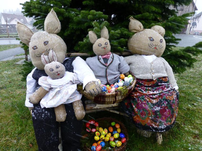 Eine Hasenfamilie mit zwei Eltern und zwei Kindern sitzt auf einer Bank vor einem Nadelbaum. Vor der Familie liegt ein Korb mit bunten Eiern.
                 title=