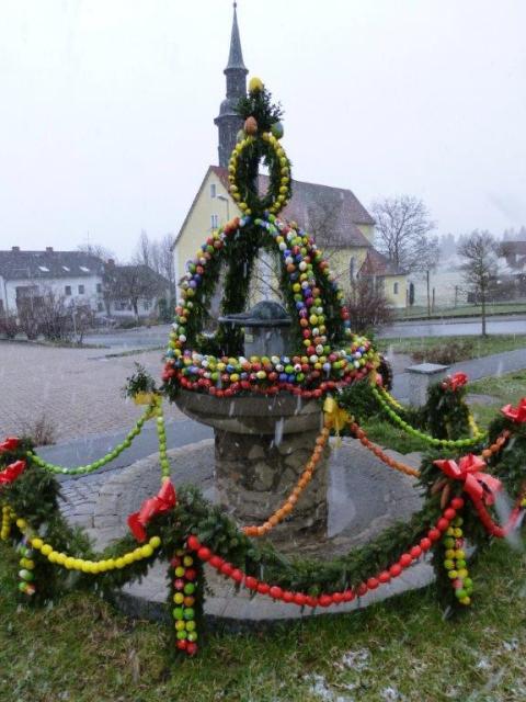 Hinter dem runden Brunnen mit Steinsäule in der Mitte, steht eine Kirche. Auf dem Brunnen steht die Krone aus grünen Girlanden mit bunten Eiern.