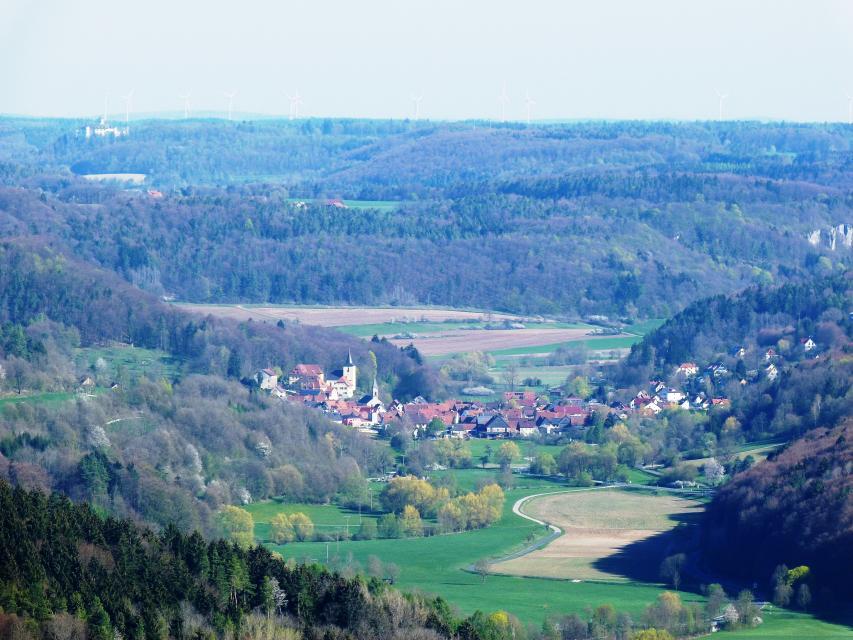 Blick von oben in ein Tal mit den Ausläufern der Hügel. Es ist in der Ferne ein Dorf mit Kirche zu erkennen.