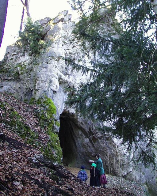 Eine Frau mit zwei Kindern steht vor dem felsigen Eingang einer Höhle im Wald.