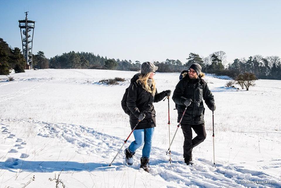 Ein Mann und eine Frau wandern mit Stöcken lächelnd durch den Schnee. Im Hintergrund erhebt sich der stählerne Aussichtsturm.
