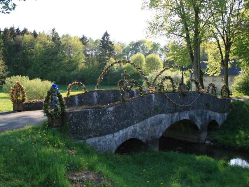 Eine alte steinerne Brücke führt über einen kleinen Bach. Die steinernen Geländer der Brücke sind mit Nadelzweiggirlanden und bunten Ostereiern geschmückt.