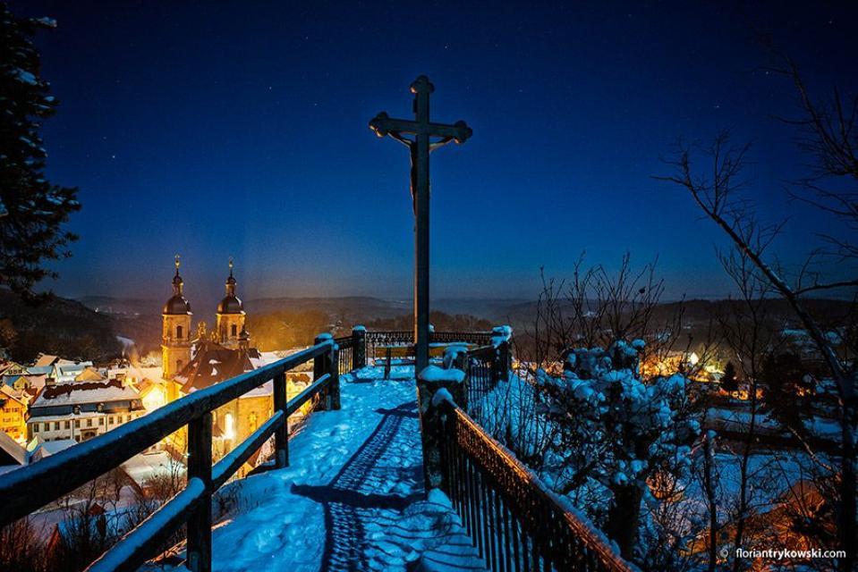 Ein verschneiter Weg mit beidseitigem Geländer führt zu einem Aussichtspunkt mit Kreuz. Unterhalb erkennt man eine mächtige Kirche.
