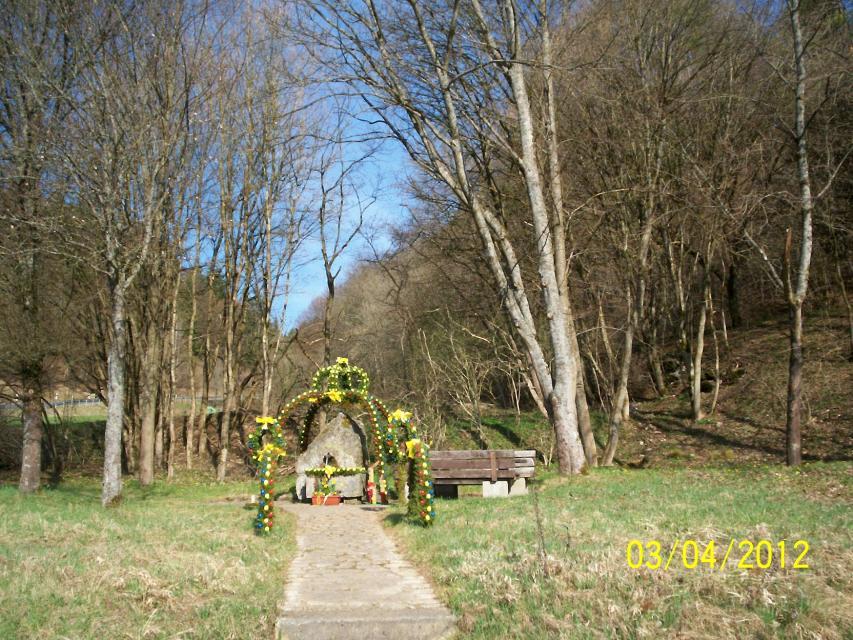 Ein Weg führt über eine Wiese zu einem mit gelben Eiern geschmückten Osterbrunnen. Im Hintergrund grenzt ein kleine Waldstück an.