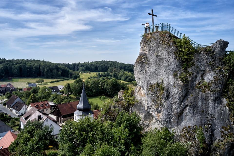Ein hochaufragender, massiver Fels mit Kreuz. Die Aussichtskanzel um das Kreuz ist eingezäunt. Besucher stehen auf dem Fels. Darunter das Dorf.