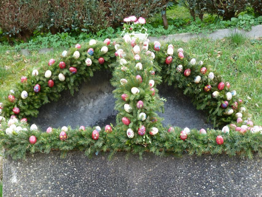 Auf einem schlichten runden Brunnen sind grüne Girlanden aus Nadelzweigen angebracht. Diese sind mit weißen und roten Ostereiern verziert.