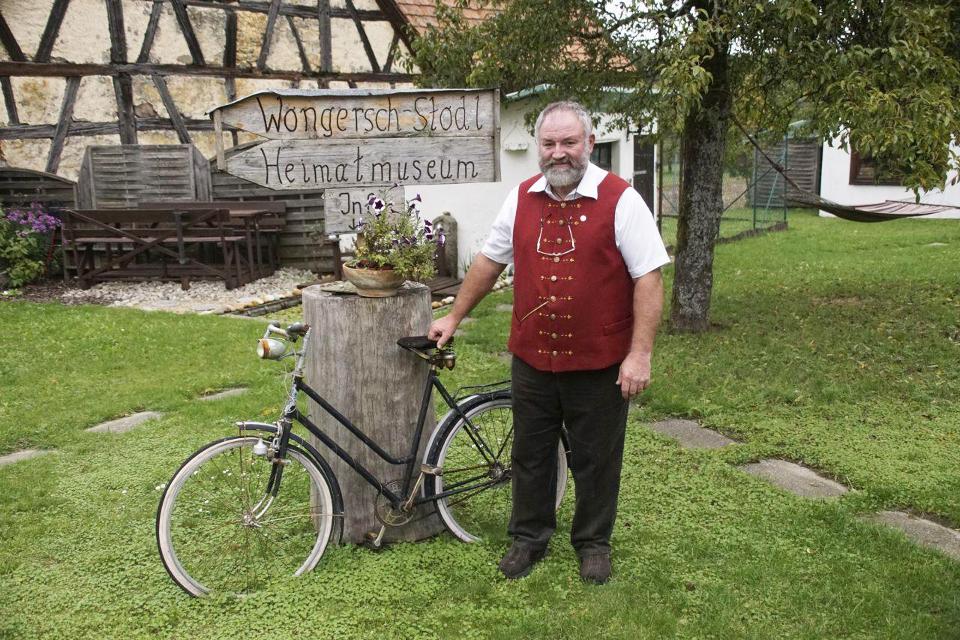 Ein Mann steht lächelnd an einem abgesägten Baumstumpf, an dem ein Fahrrad lehnt, in einer Wiese. Im Hintergrund ist eine alte Fachwerkscheune zu sehen.