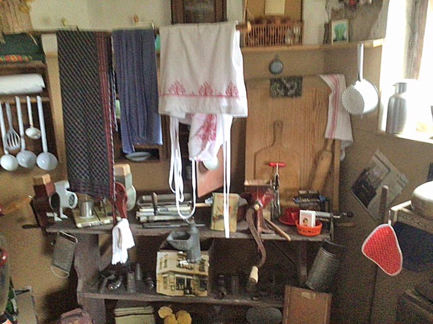 In einer Ecke sind auf einem Regal viele unterschiedliche, alte Küchenutensilien ausgestellt. Drüber hängen drei Trockentücher.