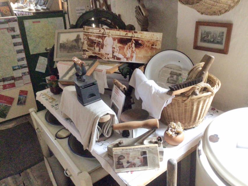 Auf einem Tisch stehen viele kleine Ausstellungsstücke. Darunter ein altes, eisernes Bügeleisen, ein geflochtener Korb und diverse alte Bilder.