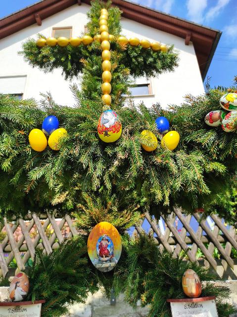 Detailaufnahme der Girlanden aus Nadelzweigen, an denen bunte Eier hängen. Darüber thront ein aus Nadelzweigen geformtes Kreuz mit gelben Eiern.
