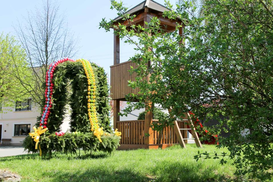 Neben einem hölzernen Spielturm, steht ein glockenförmiger Osterschmuck aus grünen Girlanden mit roten und gelben Eiern verziert.
                 title=