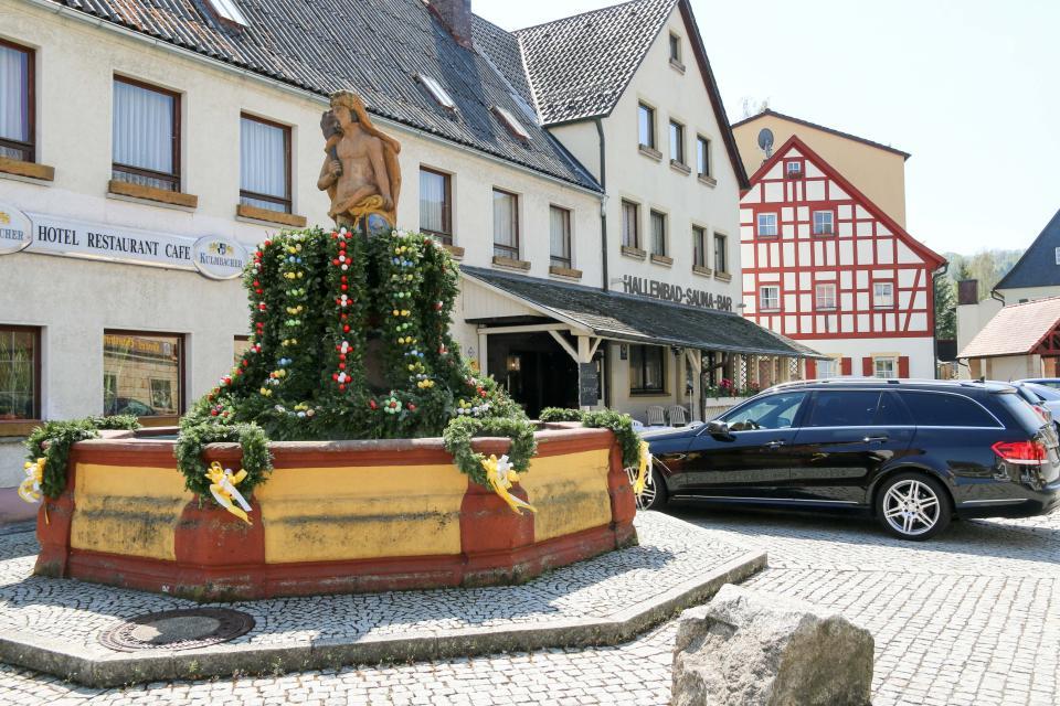 Am Marktplatz steht ein großer Brunnen, aus dessen Mitte eine Skulptur ragt. Von der Skulptur hängen grüne Girlanden mit bunten Eiern hinab.
                 title=