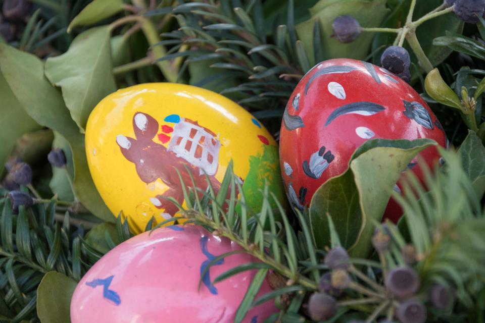 Drei Ostereier in rosa, gelb und rot zwischen Nadelzweigen. Auf dem gelben Ei ist ein Osterhase aufgemalt.