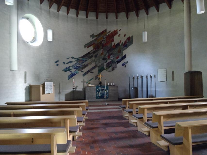 Den steinernen Altar schmückt ein Teppich, ein goldenes Kreuz und weiße Kerzen. An der Wand ist eine Mosaik Grafik in gedeckten Farben angebracht worden.
