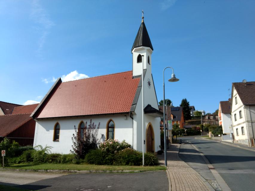 Die weiße, kleine Kapelle steht im Ortskern von Weichendorf neben einer Bushaltestelle.
                 title=