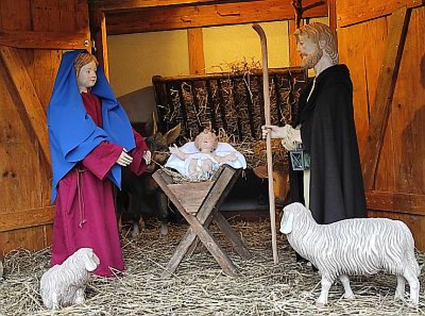 Maria und Josef stehen an der Krippe, in der das Jesuskind liegt. Davor stehen zwei Schafe.