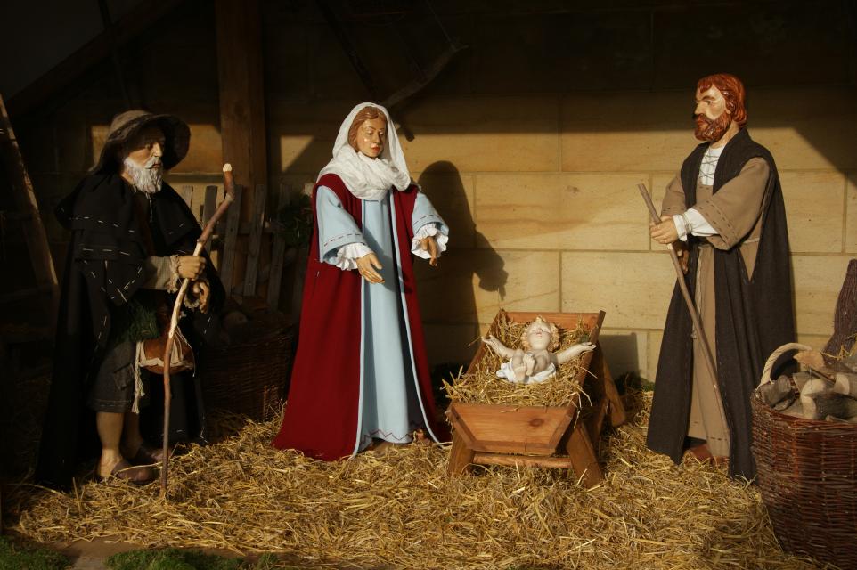 Drei mit Stoff verkleidete Krippenfiguren vor dem Kindelein, das im mit Stroh ausgelegten Trog liegt.
