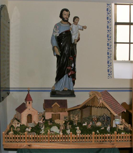 In einer Kirche steht oberhalb der Krippendarstellung mit Kapelle und Scheune eine Heiligenfigur mit einem Jungen auf dem Arm.