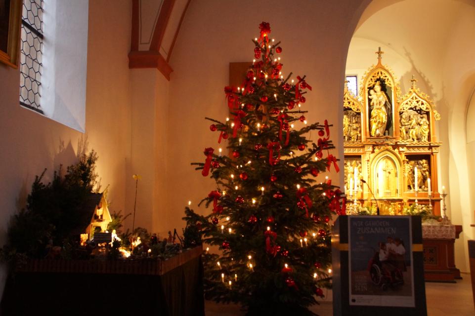 In einer Kirche steht vor dem Altar links die Krippendarstellung sowie ein beleuchteter Weihnachtsbaum.