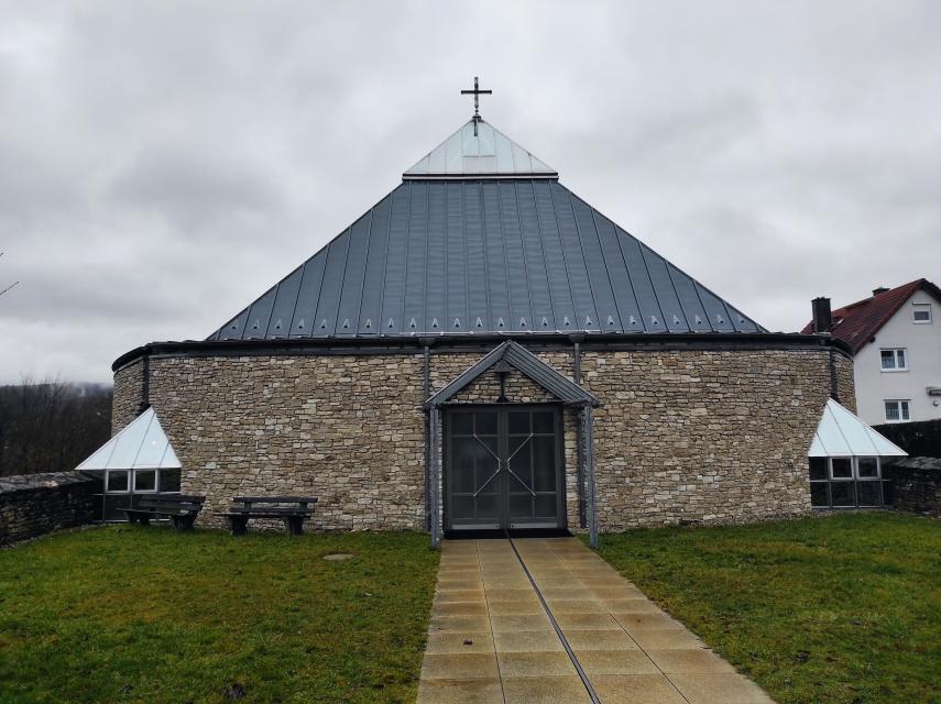 Bei bewölktem Wetter steht die aus Steinen gemauerte Kirche mit Blechdach auf der grünen Wiese am Kirchgelände. Ein Kreuz ziert die Spitze des Daches.