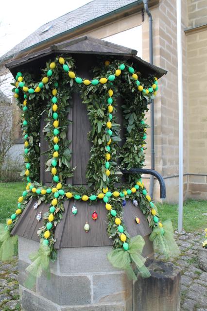 Der Brunnen, mit steinernem Sockel und hölzernen Aufbau, aus dem der Wasserspeier ragt, ist mit Girlanden und gelben und grünen Eier verziert.