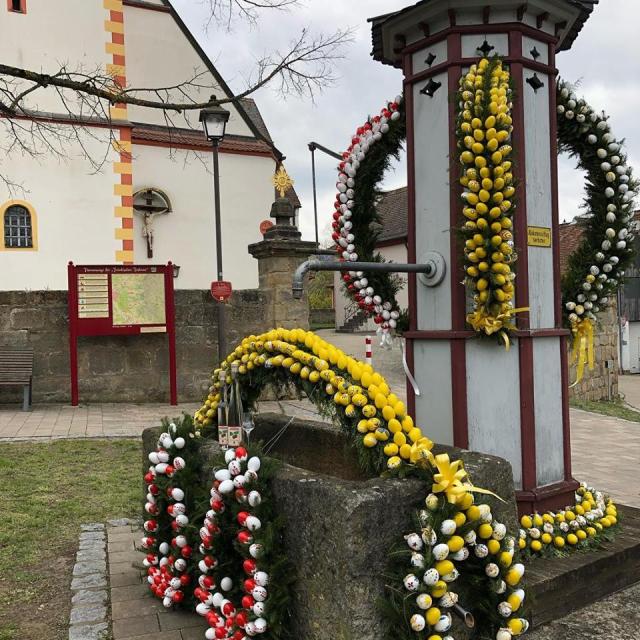 Ein Brunnen mit Steintrog sind Girlanden aus Nadelzweigen mit gelben, weißen und roten Eiern angebracht. Der Brunnen steht vor einer Kirche.
                 title=