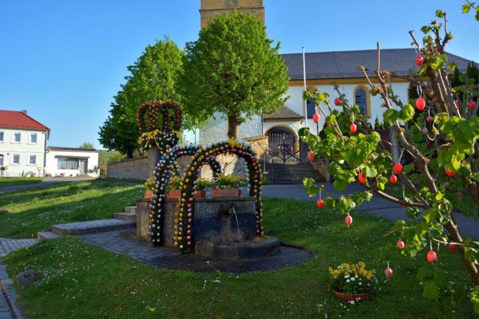 Vor der Kirche steht in einer Wiese ein Brunnen aus Steintrögen, der von mit Ostereiern geschmückten Girlanden umrahmt wird.