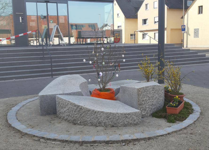 Ein moderner, runder Brunnen aus Granitsteinen, in dessen Mitte ein Blumentopf steht. An den Ästen der Pflanze hängen Ostereier.