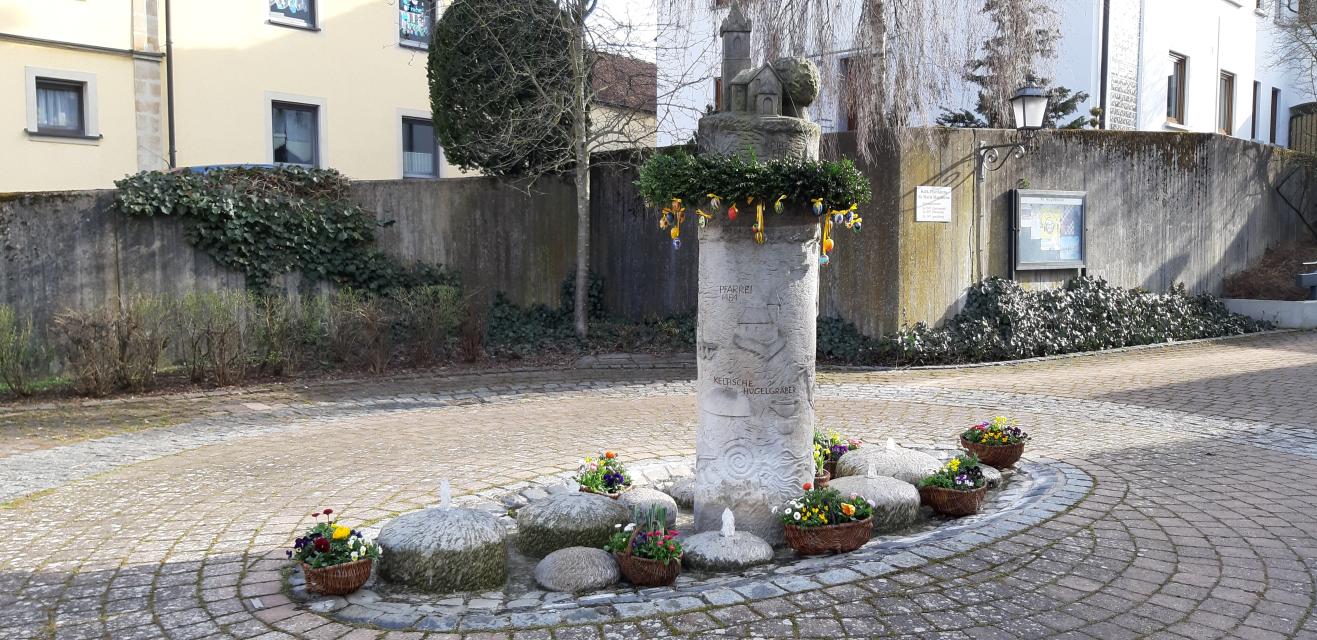 Ein Brunnen, aus einer steinernen Säule, auf der eine Kirche steht, trägt um die Säule einen grünen Kranz mit bunten Ostereiern.