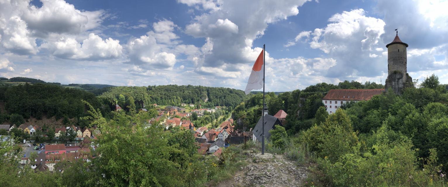 Ein Panoramabild. Im Vordergrund der Aussichtspunkt mit Fahne. Im Hintergrund erhebt sich über den Dächern des Ortes ein runder Turm.