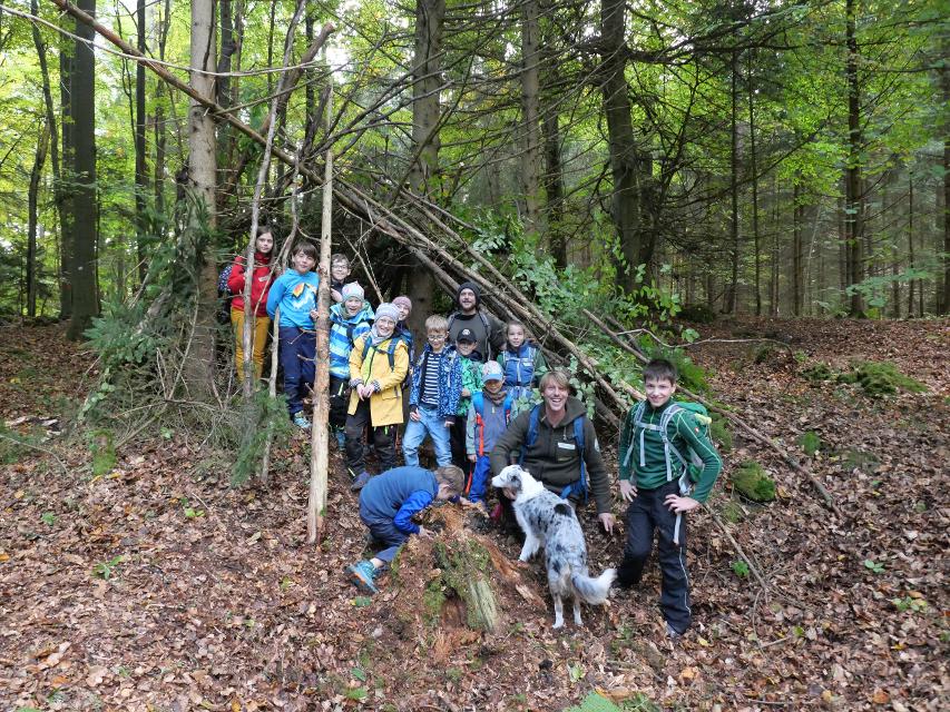 Eine Gruppe von Kindern steht im Wald in einem selbstgebautem Lager aus Ästen und Grünzeug.
                 title=