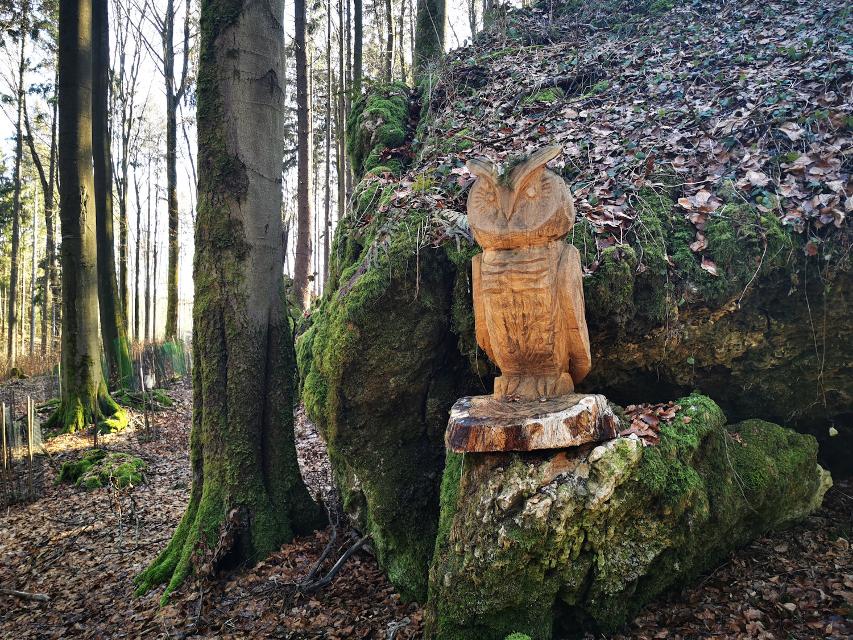 Ein aus Holz geschnitzter Uhe steht im Wald vor einem Fels.
