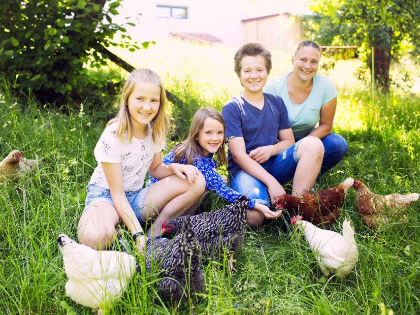 Vier Kinder sitzen in einer Wiese mit Hühnern.
                 title=