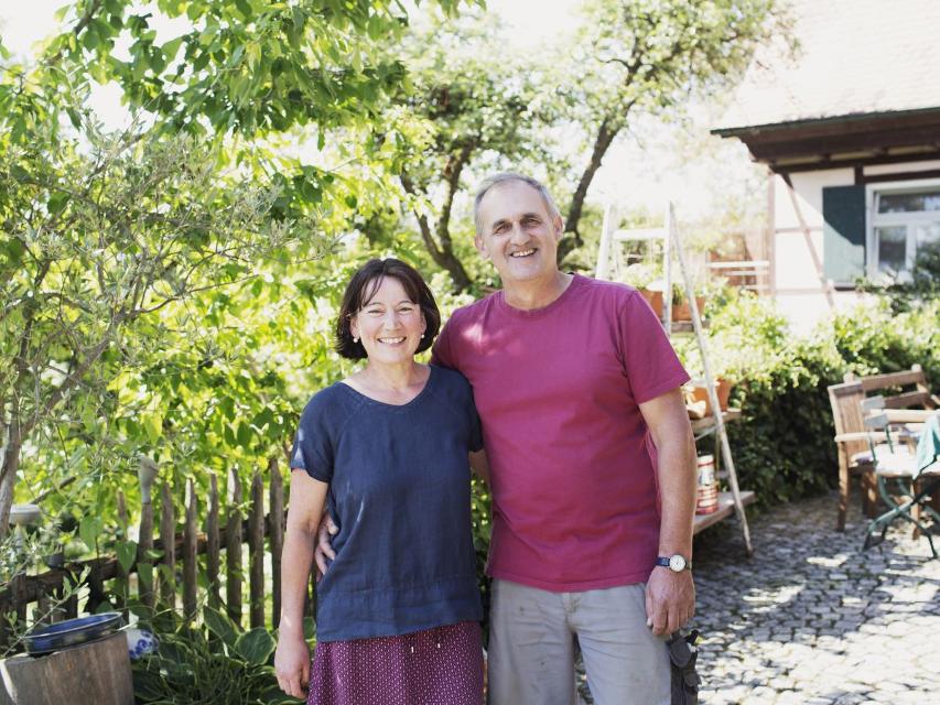 Ein Ehepaar steht Arm in Arm vor einem Zaun und Kirschbaum.
